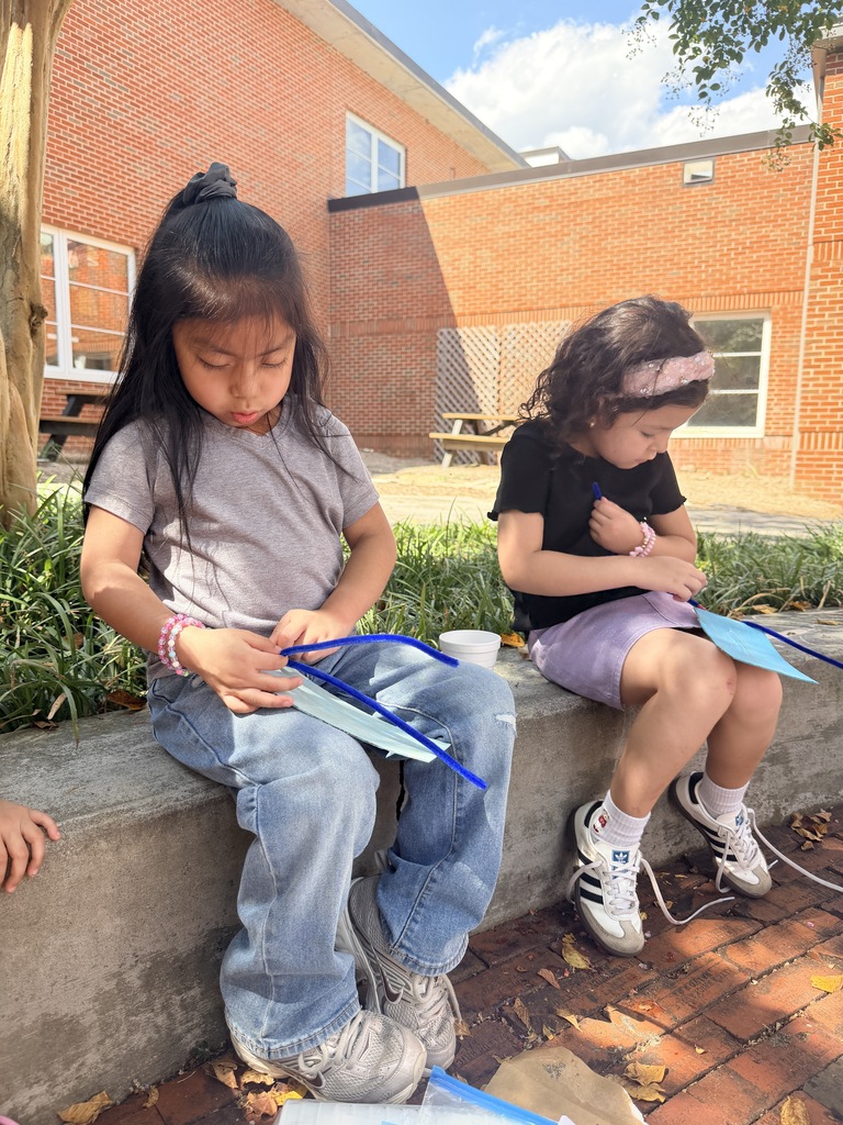 Two young girls sitting side by side on a concrete ledge outside, focused on threading beads onto blue pipe cleaners for their craft project.
