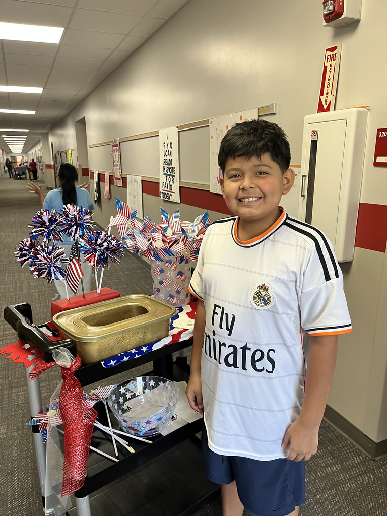 A young boy in a white Real Madrid jersey smiles while standing next to a black rolling cart decorated with red, white, and blue patriotic pom-poms, miniature American flags, and a gold ballot box for student council voting in a school hallway.
