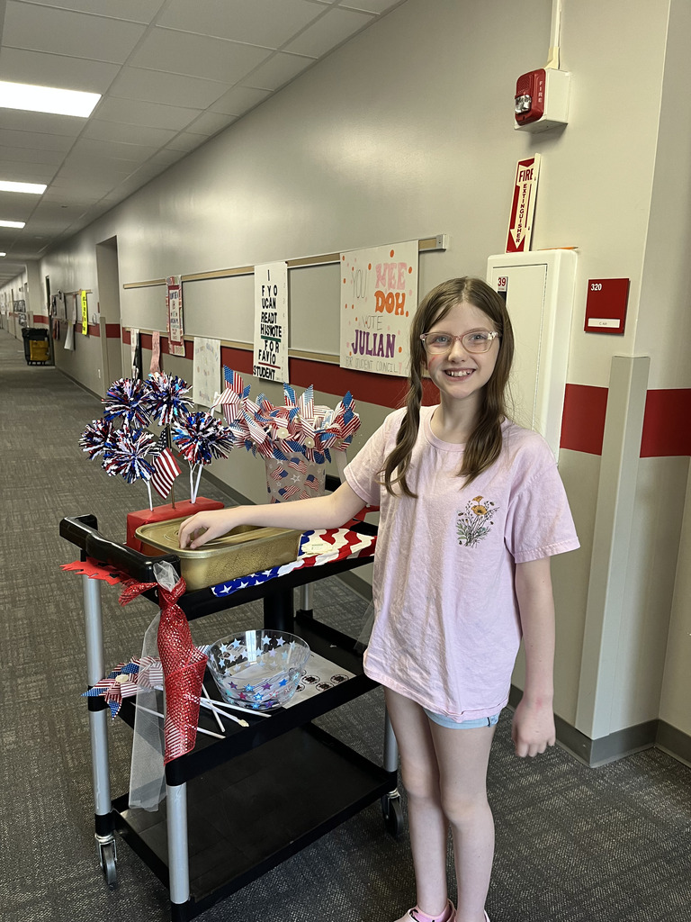 A girl wearing glasses and a light pink shirt smiles directly at the camera while placing her paper vote into a gold ballot box set on a decorated black rolling cart in a school hallway.