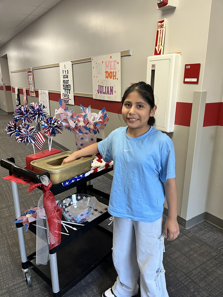 A girl in a light blue t-shirt and grey pants smiles as she reaches into a gold ballot box placed on a black rolling cart decorated with American flags, red ribbons, and patriotic poms in a school hallway lined with campaign posters.