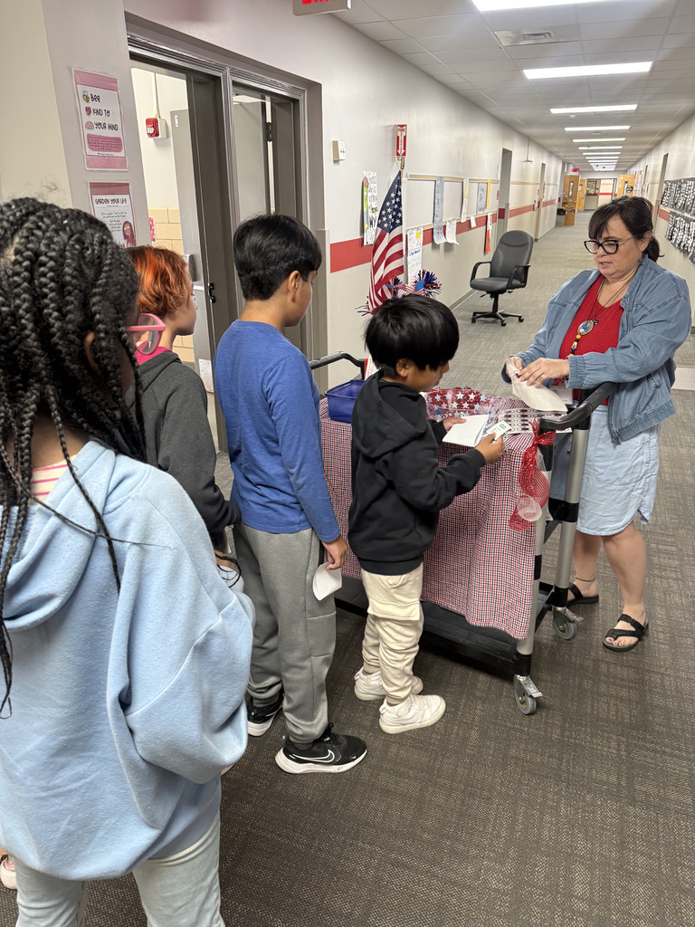 A teacher in a denim shirt assists students at a table draped in red-and-white checkered cloth and decorated with an American flag. A line of elementary students waits in the hallway to cast their votes for student council.