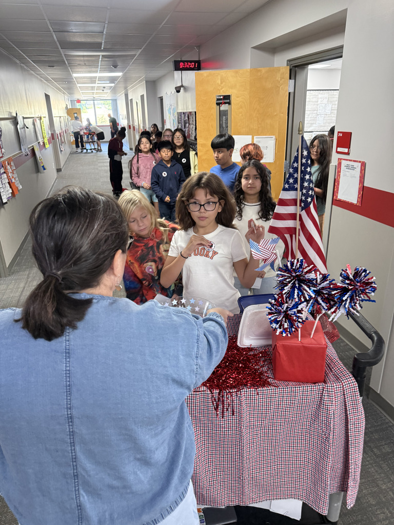 A teacher wearing glasses and a denim jacket hands paper voting slips to students lined up alongside a black rolling cart decorated with a red-and-white checkered tablecloth, patriotic ribbons, and an American flag.