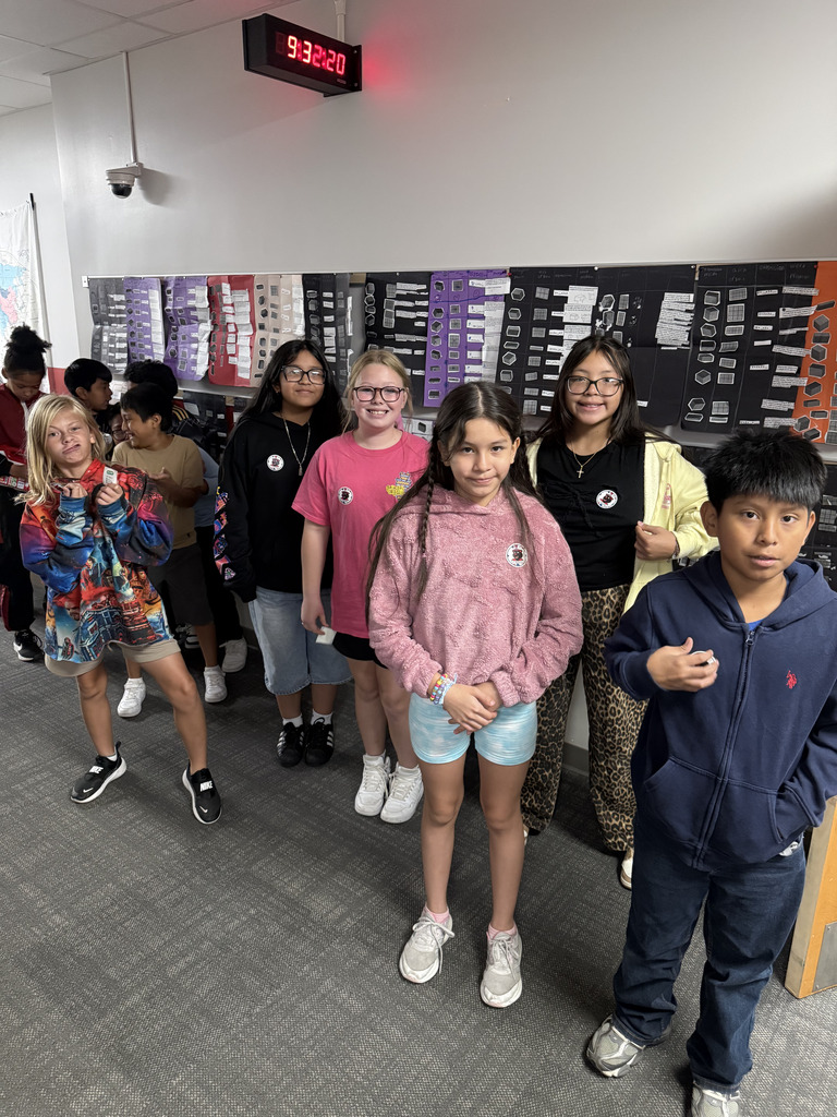 Seven elementary students pose together in a school hallway, holding small voting receipts or stickers. Student council campaign posters and classroom displays line the wall behind them.