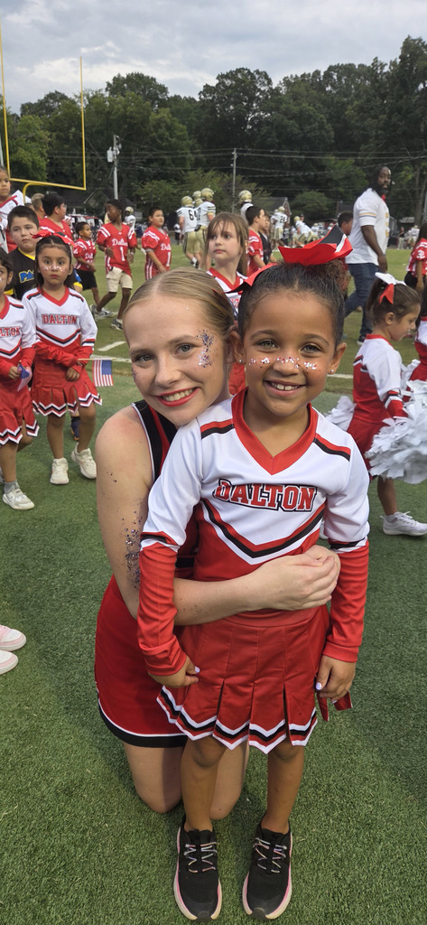 A high school cheerleader wearing a red, white, and black "DALTON" uniform kneels on a green football field, smiling and wrapping her arm around a young elementary girl dressed in a matching youth cheerleading outfit. Both have glitter painted on their cheeks, and other youth cheerleaders and football players are visible in the background.