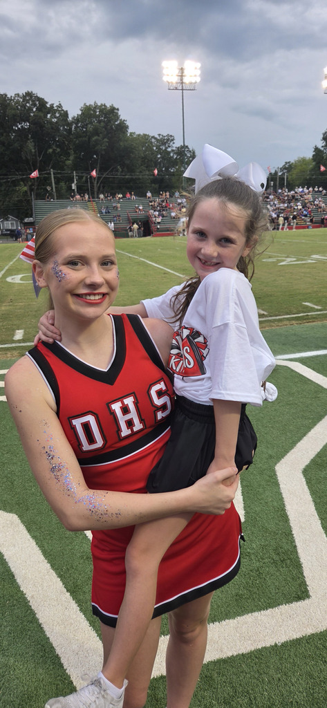 A high school cheerleader in a red "DHS" uniform holds a young girl wearing a white "KITTENS" cheer shirt and a white hair bow in her arms. They both smile at the camera on a football field, with stadium bleachers and field lights in the background under an overcast sky.