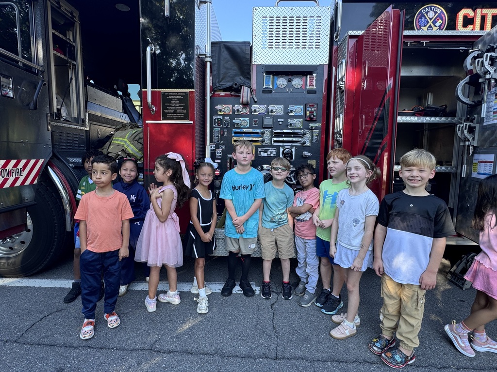 A line of elementary students stands outdoors in front of a red fire truck, smiling for a group photo during a school field trip or demonstration. Open compartments and equipment panels on the side of the fire truck are visible behind them.