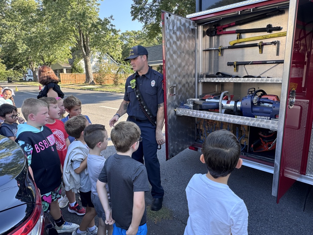 A firefighter in a navy uniform gestures as he shows rescue tools, including hydraulic spreaders and axes stored inside an open compartment of a red fire truck, to a group of interested elementary students standing nearby.