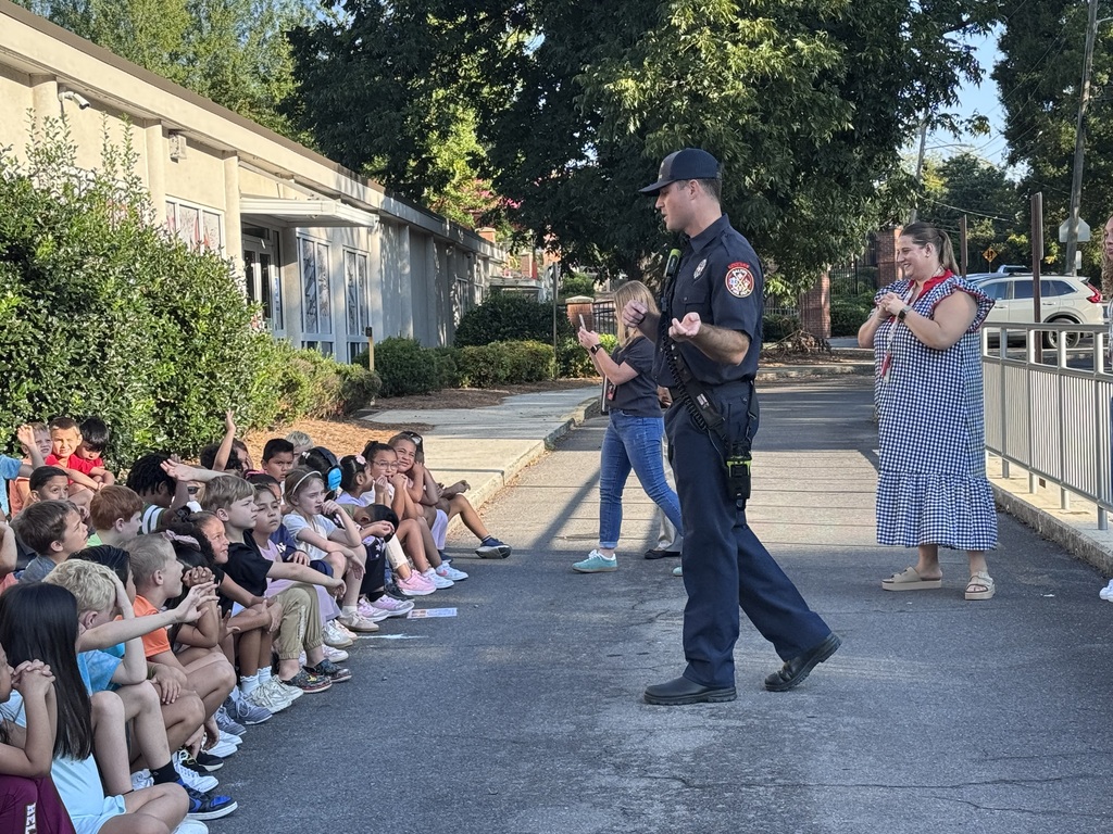 A firefighter in a dark navy uniform and cap speaks gestures while walking past a group of elementary students sitting along a sidewalk. Teachers and staff members stand nearby, smiling and watching the presentation outside a school building.