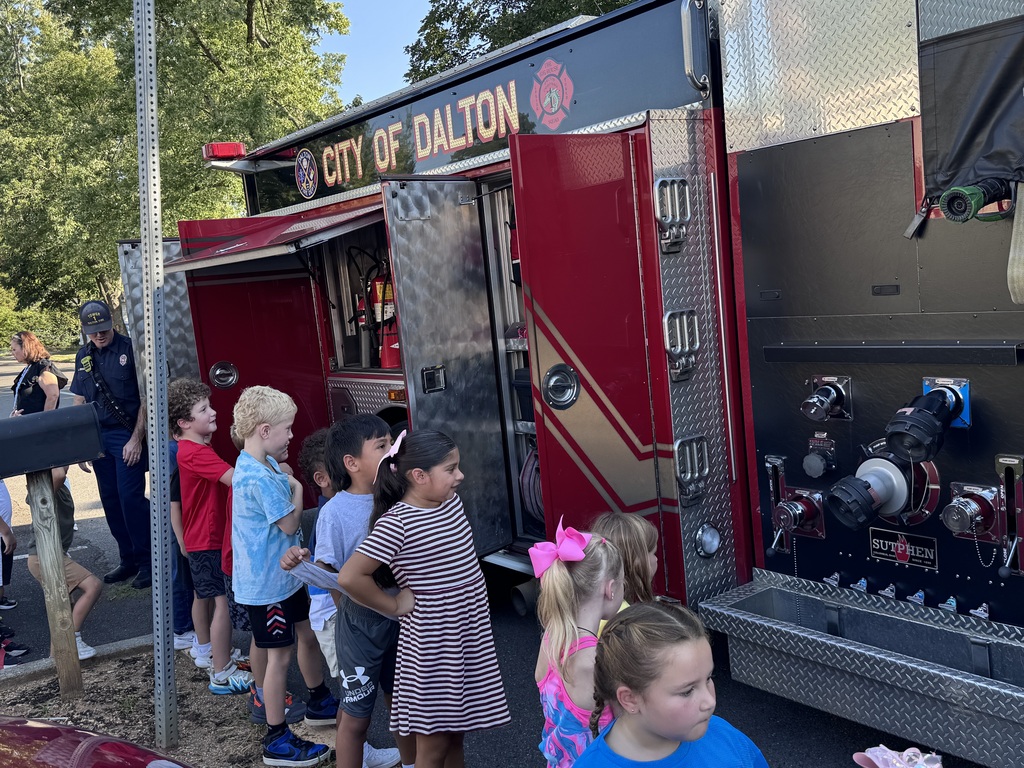 A group of elementary students line up beside a red City of Dalton fire truck to look at the hose connections and open storage compartments, with a firefighter standing nearby in the background.