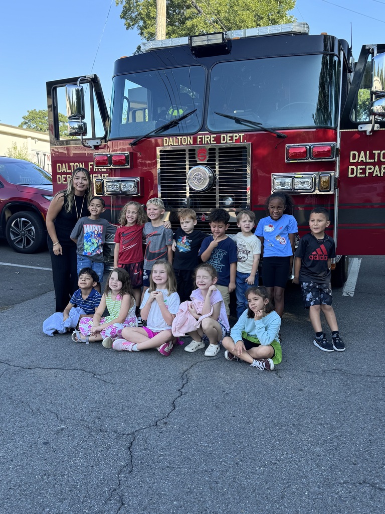 A group of elementary school students poses in front of a red Dalton Fire Department truck alongside a teacher, with some sitting on the ground and others standing in a row in front of the vehicle's grill.