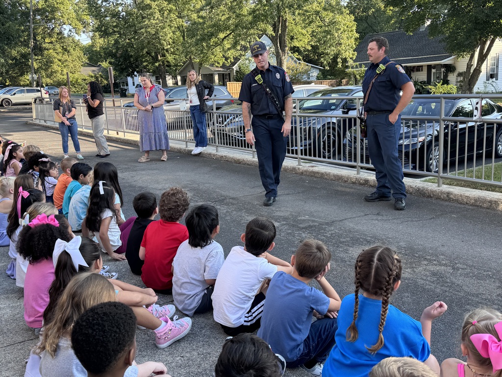 Two firefighters in dark blue uniforms stand outdoors on a paved area, speaking to a group of young elementary students sitting on the ground listening. Several school staff members stand along a railing in the background watching.