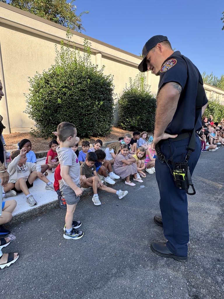 A firefighter in a navy uniform bends down slightly to speak one-on-one with a young elementary student standing in front of him outside a school. Other students sit nearby along a curb watching the interaction.