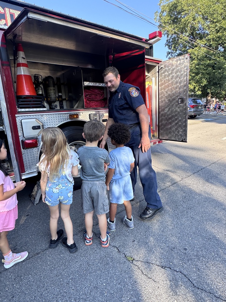 A smiling firefighter in a navy uniform stands beside an open storage compartment of a red fire truck, showing equipment to a small group of young elementary students.