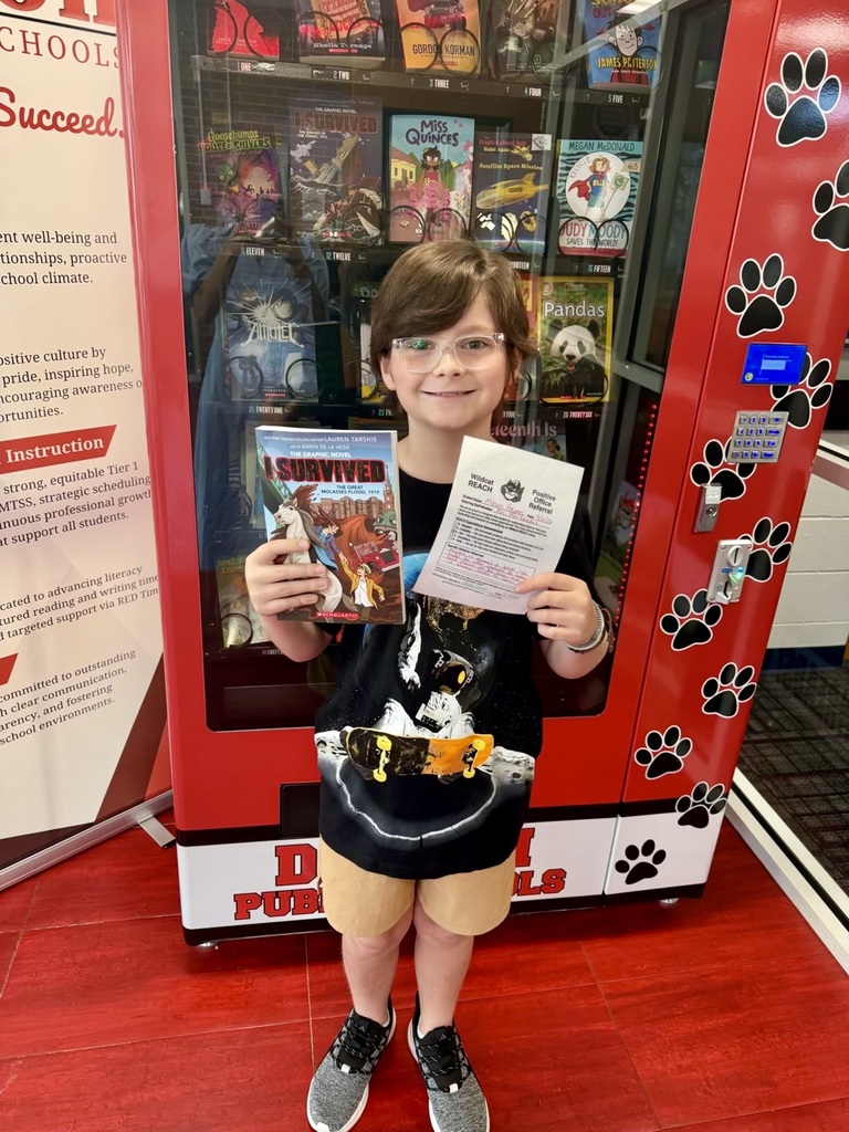 A young student wearing clear glasses, a graphic t-shirt, and beige shorts stands smiling in front of a red book vending machine. He holds a graphic novel titled I Survived The Great Molasses Flood, 1919 in one hand and a Wildcat REACH Positive Office Referral certificate in the other. The book vending machine behind him is filled with popular children's books and decorated with black paw prints.