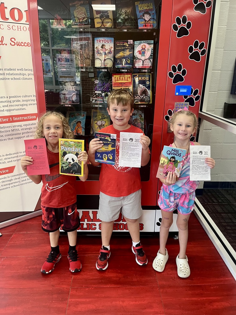 Three young students stand side-by-side smiling in front of a red Dalton Public Schools book vending machine decorated with black paw prints.  The child on the left, wearing a red Nike shirt and patterned athletic shorts, holds up a National Geographic Kids book about pandas alongside a pink Positive Office Referral certificate.  The child in the center, wearing a bright red shirt and gray shorts, holds a Magic School Bus Rides Again book along with his referral form.  The girl on the right, wearing a colorful, swirling pink-and-blue outfit with white clogs, holds up the book Miss Quinces and a white referral certificate decorated with hearts.
