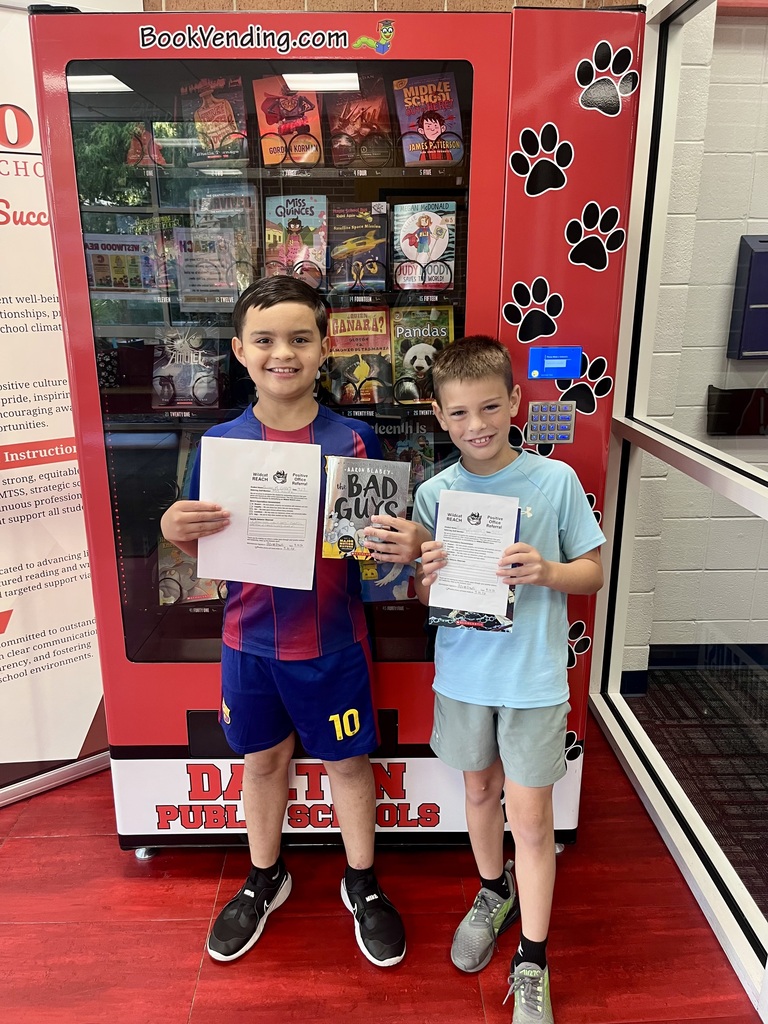 Two young students stand side-by-side smiling in front of a red Dalton Public Schools book vending machine adorned with black paw prints. The student on the left wears a blue and red soccer jersey matching his shorts and holds up a Wildcat REACH Positive Office Referral certificate alongside the book The Bad Guys by Aaron Blabey. The student on the right wears a light blue athletic shirt and gray shorts while holding his own Positive Office Referral certificate and a book.