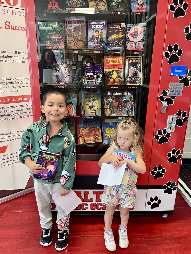 Two young students stand side-by-side in front of a red Dalton Public Schools book vending machine decorated with black paw prints.  The boy on the left wears a light green patterned pullover sweater, gray sweatpants, and black sneakers. He smiles as he holds a Goosebumps House of Shivers book and a paper certificate.  The girl on the right, wearing a floral overall outfit and white sneakers, holds the book The Tryout along with her referral paper.