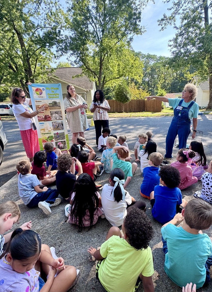 A group of elementary school students sits outdoors on a paved surface listening to an educational presentation on nutrition and agriculture. On the left, an instructor holds up a poster titled "My Plate Is Georgia Grown," which breaks down food categories like grains, protein, dairy, and vegetables. To the right, another presenter in denim overalls points toward the audience, guiding the interactive lesson while other staff members observe.