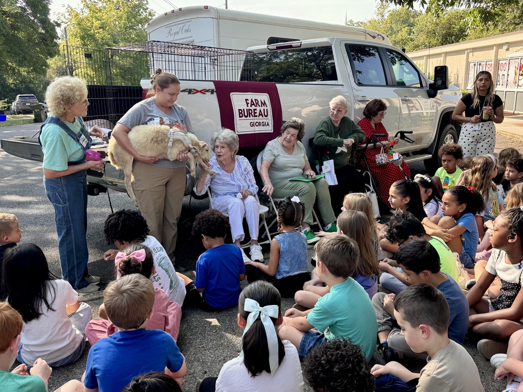 An instructor holds a small lamb for a seated senior guest to pet during an outdoor agricultural demonstration for elementary students. The children sit in a group on the pavement watching the interaction in front of a white pickup truck bearing a "Georgia Farm Bureau" banner on its tailgate.