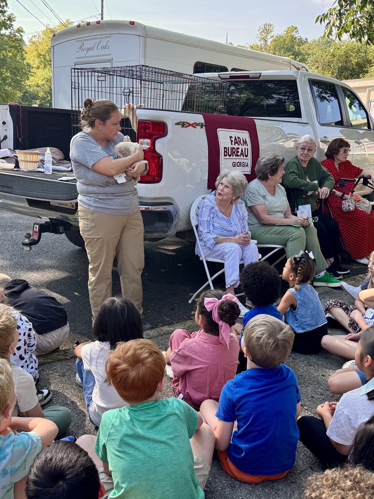 A female instructor stands in front of a group of elementary students sitting outdoors on a paved surface, holding a small white chicken during an agricultural demonstration. Behind her is a white pickup truck displaying a "Georgia Farm Bureau" banner on the tailgate and carrying a wire animal crate. To the side, several seated adult visitors watch the presentation while the children face forward, attentive to the speaker.