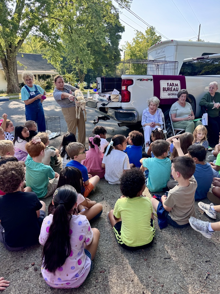 A group of young elementary students sits on an outdoor paved driveway listening to an educational agricultural demonstration. In the background, a presenter holds a small lamb near the open tailgate of a white pickup truck bearing a "Georgia Farm Bureau" banner. Adults and community volunteers, including a woman in denim overalls and seated senior guests, look on as the children watch the presentation.