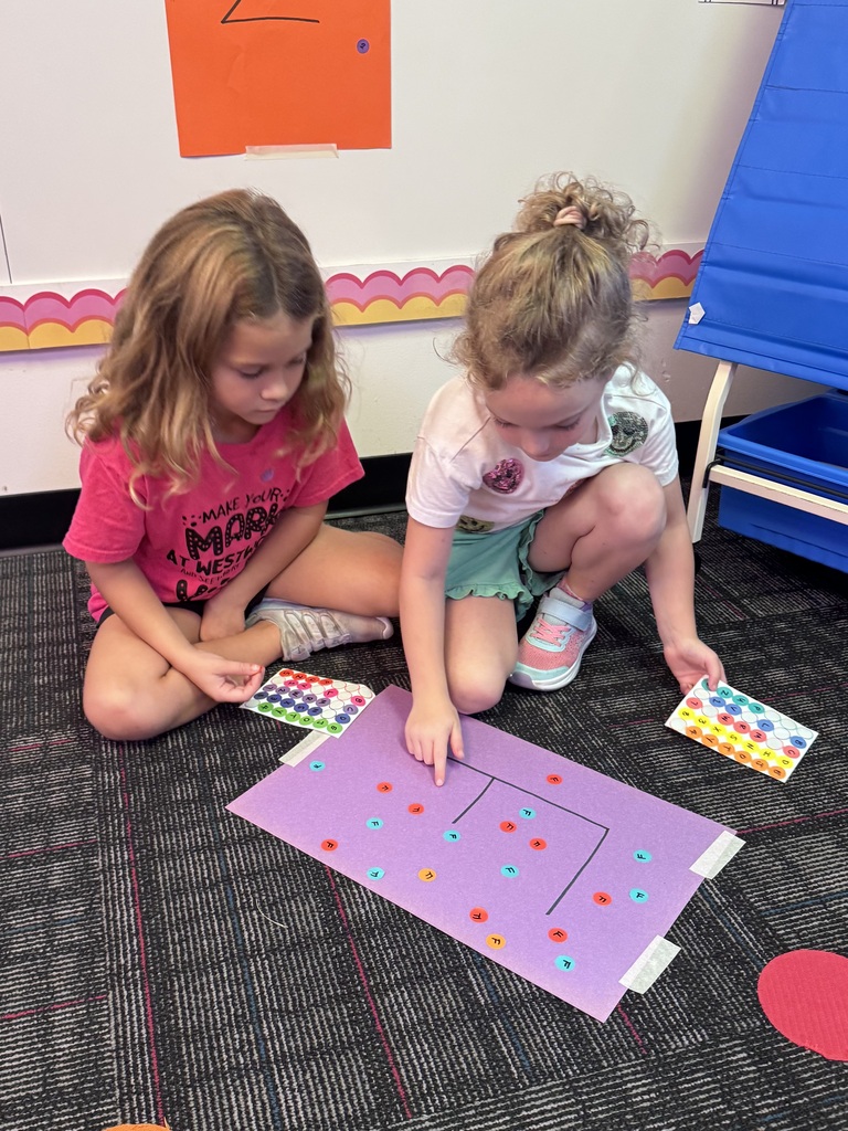 Two young girls sit on the classroom rug working together on a Dot Day activity. One points to colored dot stickers placed on a purple construction paper chart while using sticker sheets