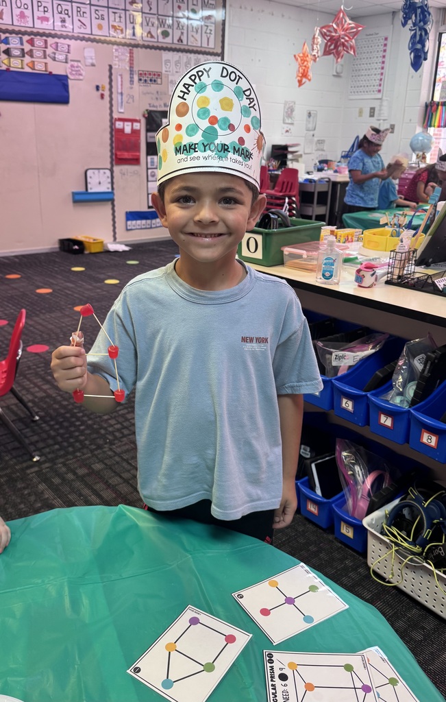 A young student wearing a decorated "Happy Dot Day" paper crown proudly holds up a geometric structure built with toothpicks and candy dots while standing in front of instructional activity cards.