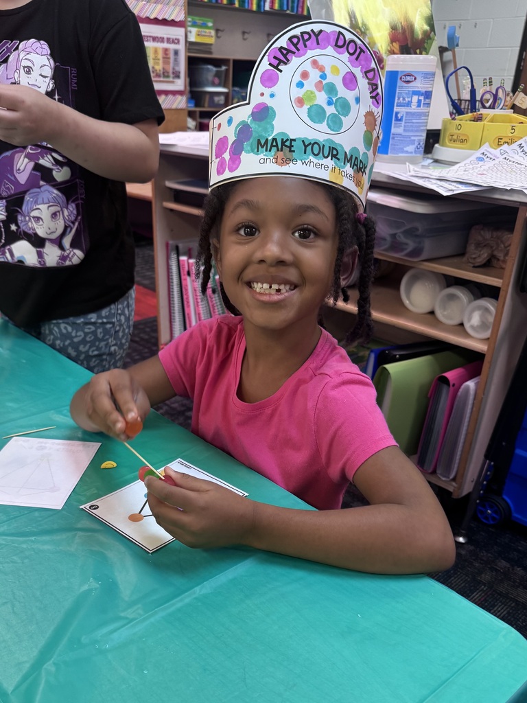 A young girl smiling and wearing a decorated paper "Happy Dot Day" crown sits at a table building a 3D shape using toothpicks and candy dots.