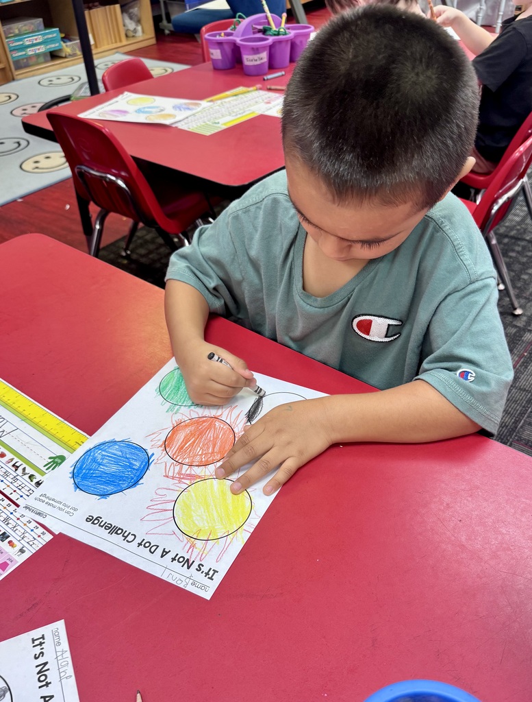 A young student works attentively at a red table, using a black crayon to transform colorful drawn circles on an "It's Not A Dot Challenge" activity page.