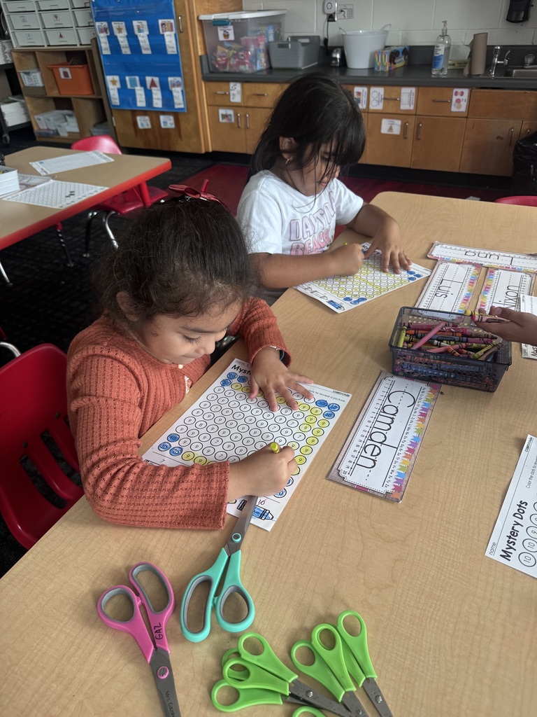 Two young students sit at a classroom table using yellow crayons to complete "Mystery Dots" color-by-number activity sheets near a basket of crayons and child-safe scissors.