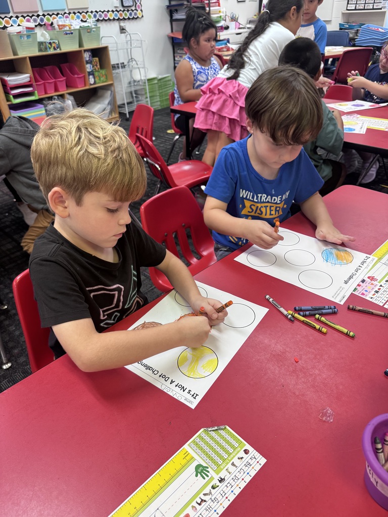Two students sit at a red classroom table using crayons to color and transform large circles on "It's Not A Dot Challenge" worksheets.