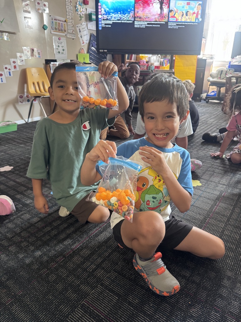 Two young boys sit on the classroom floor smiling while holding up labeled clear plastic bags filled with colorful Dot Day snack mixes.