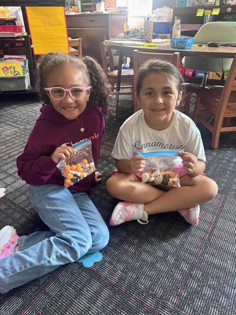 Two young girls sit cross-legged on the carpet holding up clear plastic bags containing Dot Day snack mixes featuring round cereal pieces and treats.