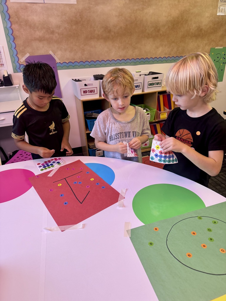 Three young students gather around a table peeling and applying colorful dot stickers onto letter activity sheets and colored paper during a classroom activity.
