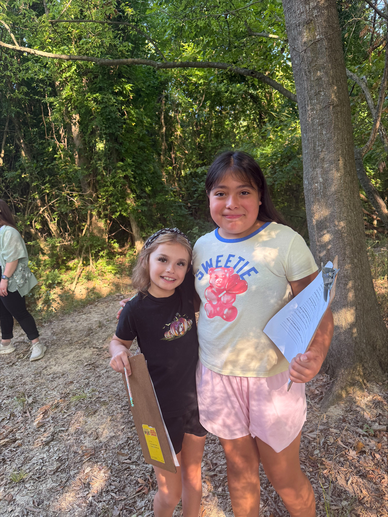 Two young girls pose arm-in-arm along a dirt path framed by trees. The girl on the left wears a black shirt with a pumpkin print, while the girl on the right wears a graphic tee with a gummy bear that says "Sweetie." Both hold clipboards with activity sheets during an outdoor lesson.