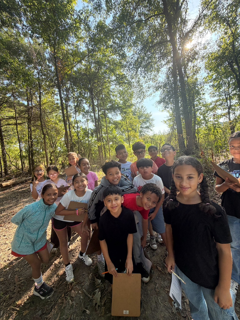 A large group of elementary school children gather closely together on a dirt path under the canopy of trees. They look up and smile at the camera, with many holding clipboards, pencils, and worksheet papers.