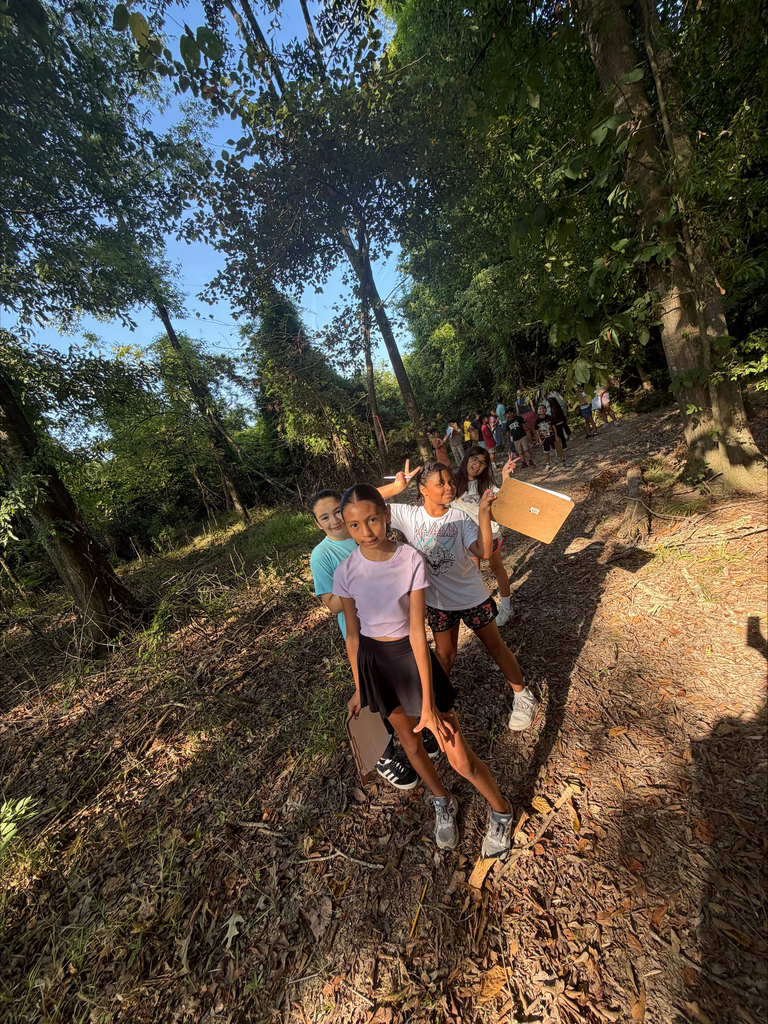 Four elementary school girls stand on a fallen leaf-covered trail surrounded by trees. The girl in front wears a purple top and black skirt while posing with a clipboard, as her friends behind her make peace signs and smile for the camera.