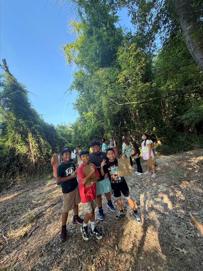 A small group of elementary students stand on a sunlit dirt path surrounded by tall green trees. Four boys pose together in the foreground holding wooden clipboards, while other classmates walk along the woodland trail in the background.