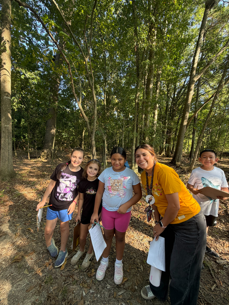 A teacher wearing a yellow shirt stands alongside four students holding clipboards in a dense forest setting. They all smile toward the camera during an outdoor learning activity amidst the trees.