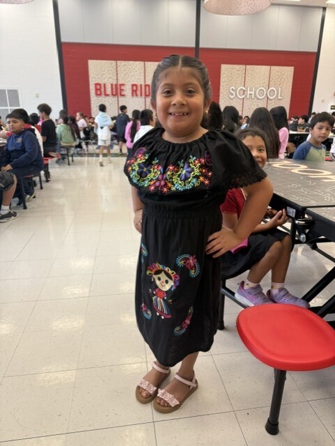 Main Subject: A young girl smiles while standing in the center of the frame, posing with one hand on her hip. She is wearing a traditional black off-the-shoulder dress decorated with colorful floral embroidery around the neckline and an embroidered doll design on the skirt. Background & Setting: The photo is taken inside the Blue Ridge School cafeteria, with large red signs reading "BLUE RIDGE" and "SCHOOL" visible on the wall behind her. Other elementary students are sitting at cafeteria tables in the background.
