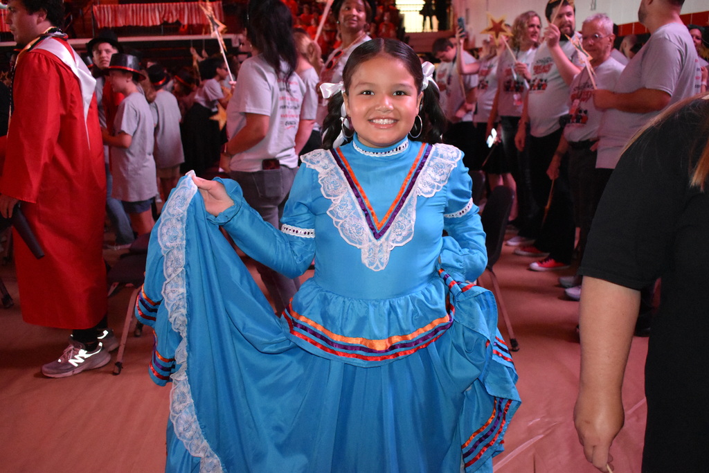 Main Subject: A young girl smiles directly at the camera, holding out the side of her long, bright blue traditional dress. The dress features long sleeves, white lace trim along the neck and hem, and colorful ribbon accents along the collar and skirt.  Background & Setting: The photo is taken at a crowded indoor event, likely a school assembly or parade. In the background, adults and students in red gowns and grey t-shirts are standing and walking through the room.