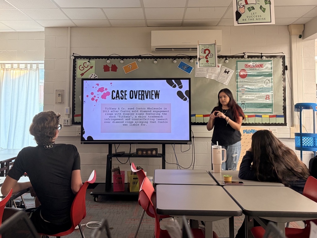A student giving an oral presentation in a classroom using a digital screen display.  Presenter: A female student stands to the right of a large screen, speaking and gesturing with her hands, wearing a black shirt and jeans with a large insulated tumbler on the desk nearby.  Digital Display: A TV monitor displays a slide titled "CASE OVERVIEW" in bold lettering. The text box details a legal trademark case between Tiffany & Co. and Costco Wholesale.  Audience: Two students are visible in the foreground seated at grey school desks, looking forward at the presentation.  Classroom Environment: The room features white brick walls decorated with educational posters, including Monopoly-themed bulletin board decorations above the main screen.