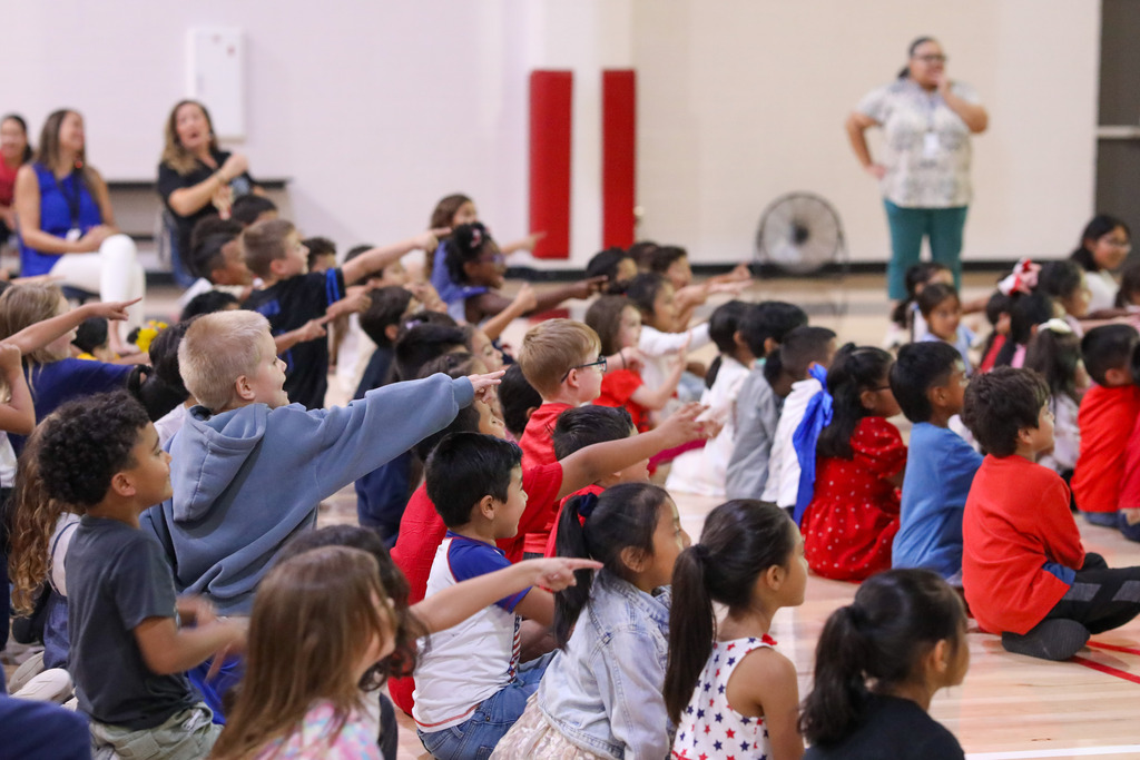 Young elementary students sitting on the gymnasium floor watching a stage performance.