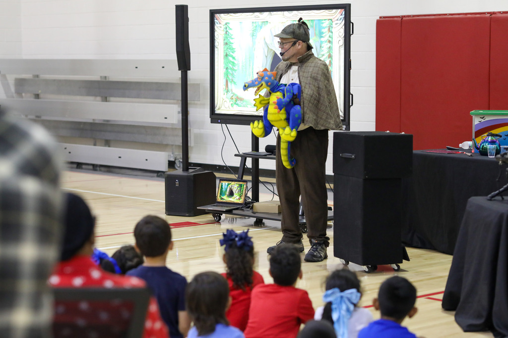 A presenter holding a colorful dragon puppet in front of seated elementary students during an assembly.