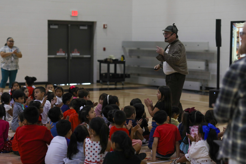 Young elementary students sitting on the gymnasium floor watching a stage performance.
