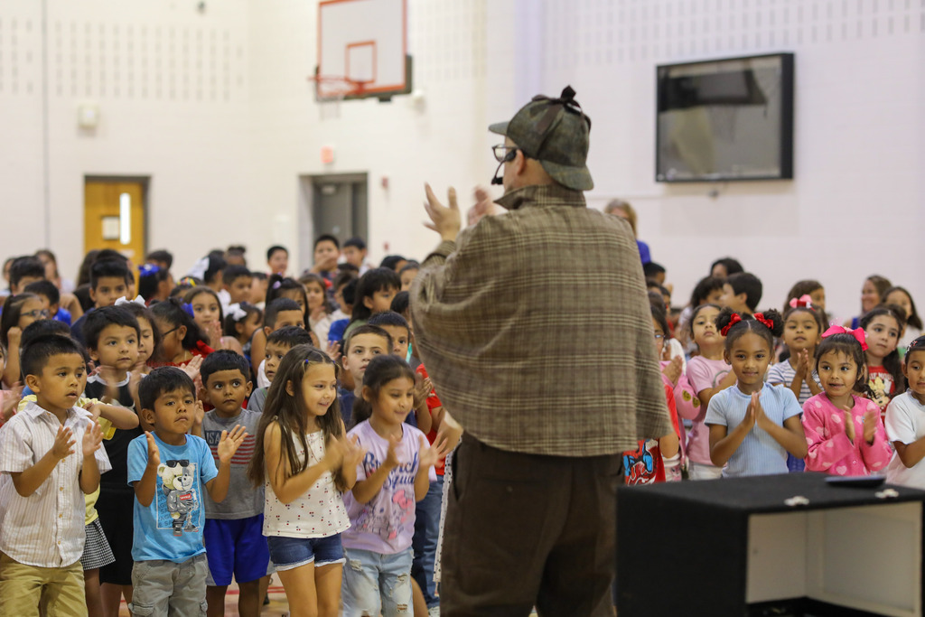 A performer in a detective costume presenting to a room full of clapping elementary students.