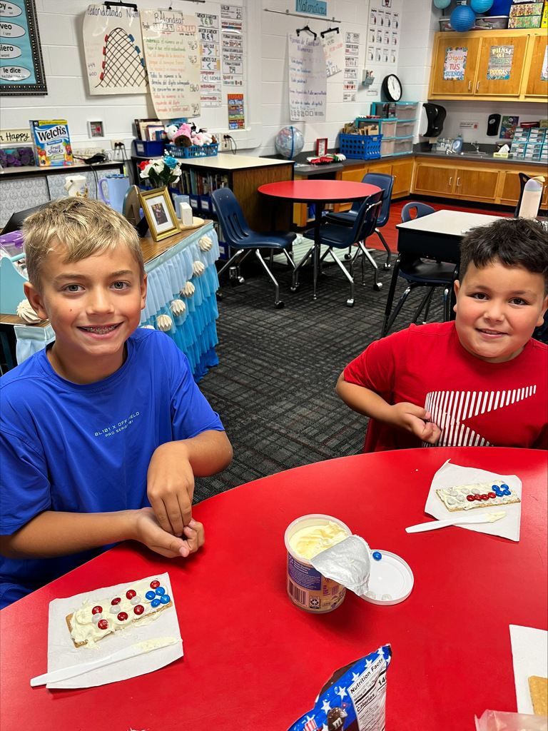 Two elementary students smile at a red classroom table while making American flag graham cracker treats using white frosting and red and blue candy-coated chocolates.