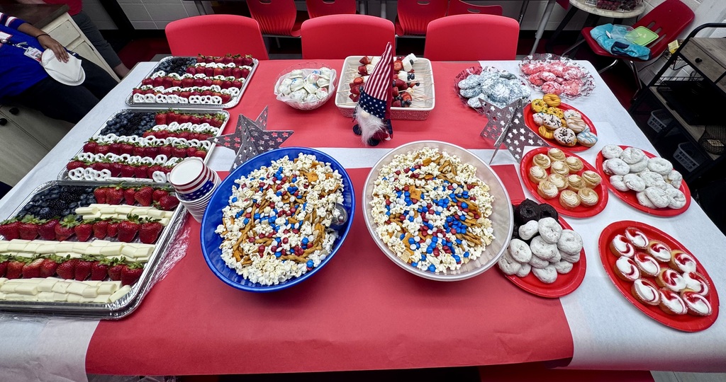 A festive buffet table covered in red and white tablecloths is filled with red, white, and blue patriotic treats. The spread includes fruit platters arranged like American flags, large bowls of snack mix, frosted donuts, cookies, and small patriotic decorations.