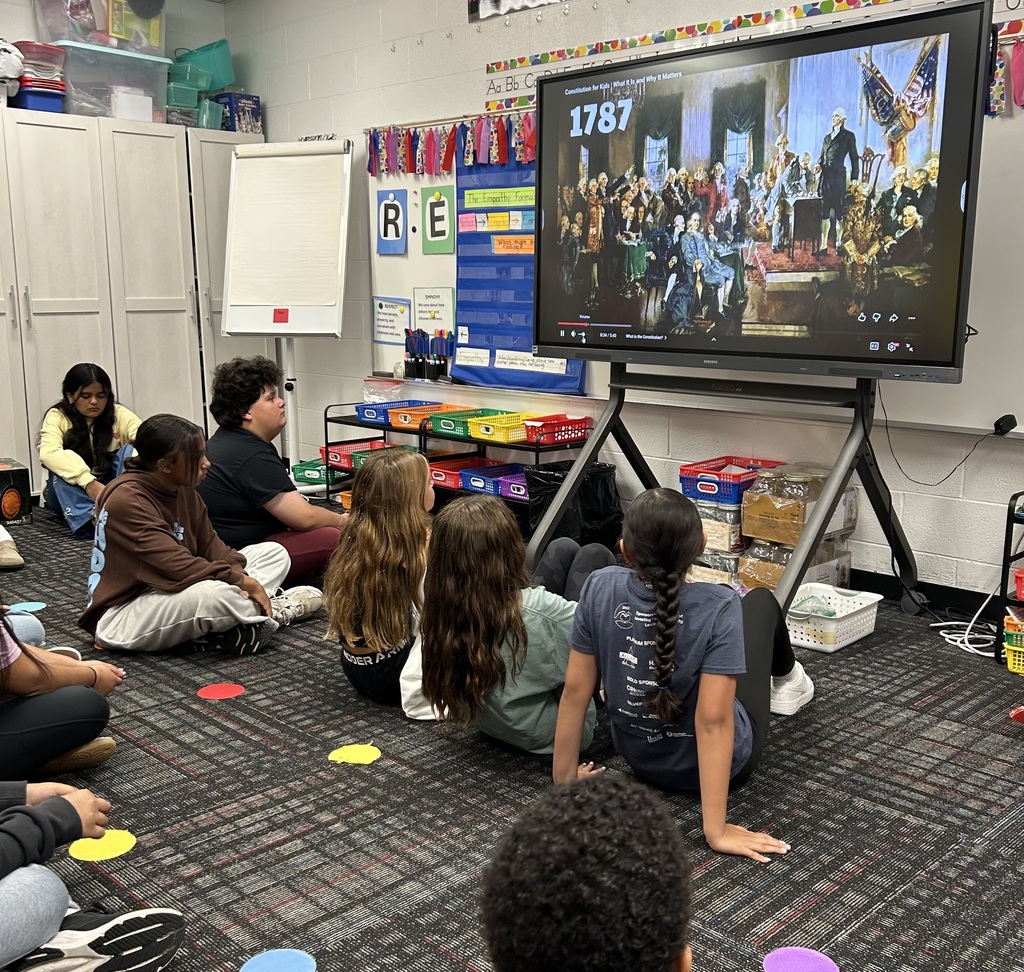 Elementary students sit cross-legged on a classroom rug, watching an educational video about the 1787 Constitutional Convention on a large smart display board. Colorful floor markers and organized classroom storage bins frame the learning space.
