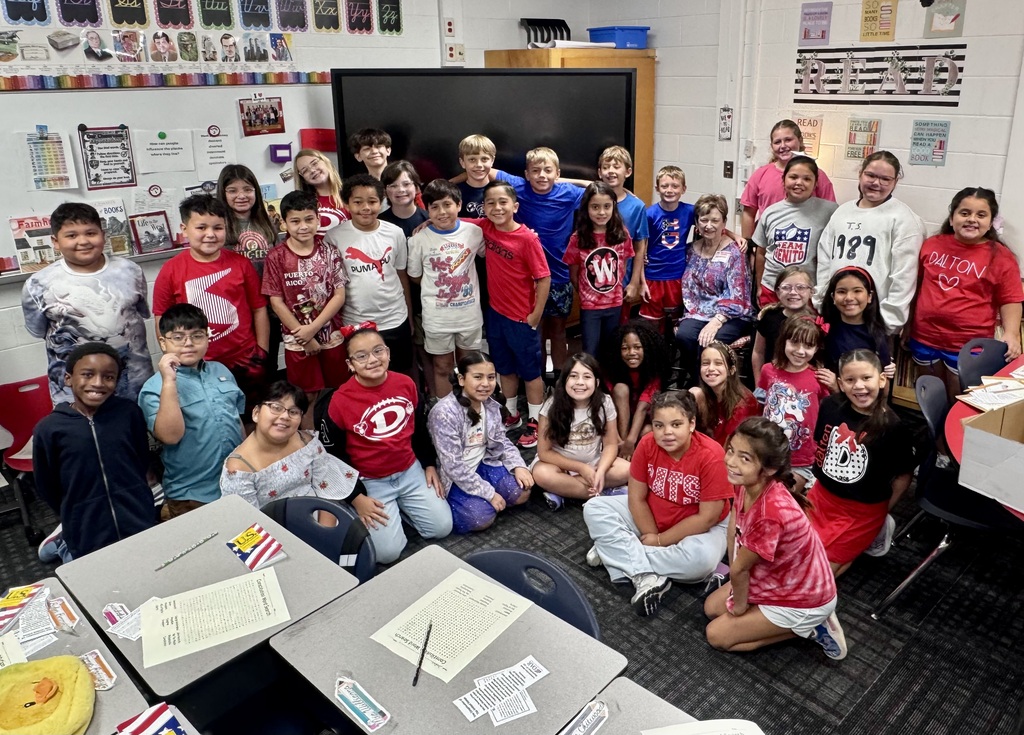 A diverse group of elementary school students and adults pose together for a group photo inside a colorful classroom. Some students are standing in the back in front of an interactive display board, while others sit on the floor in the front. Worksheets with pencil activities and small American flag markers rest on student desks in the foreground.